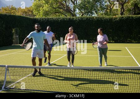 Divers amis en vêtements de sport marchant ensemble sur un court d'herbe portant des raquettes de tennis à côté du filet Banque D'Images