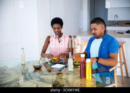 Couple diversifié assis à la table à manger en verre dans la cuisine à la maison manger des hamburgers avec ketchup, moutarde Banque D'Images