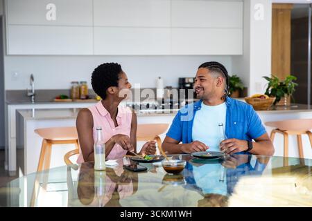 Couple diversifié assis à la table en verre dans la cuisine manger salade avec smartphone, bouteilles en verre Banque D'Images