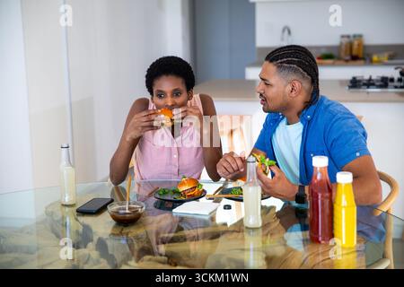 Couple diversifié mangeant des hamburgers, salades d'accompagnement à la table à manger en verre avec bouteilles de condiment dans la cuisine Banque D'Images