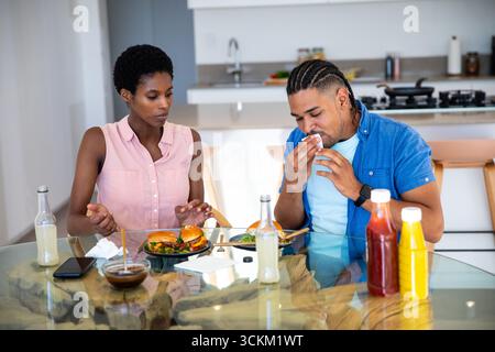 Couple diversifié mangeant des hamburgers et de la salade à la table à manger en verre dans la cuisine moderne avec des condiments Banque D'Images
