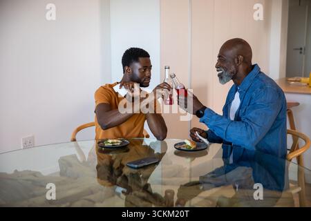 Père et fils afro-américains serrant des bouteilles de boisson rouges à table avec smartphone Banque D'Images