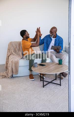 Père afro-américain et son fils haut-fiving sur un canapé léger dans le salon avec tablette, tasses Banque D'Images