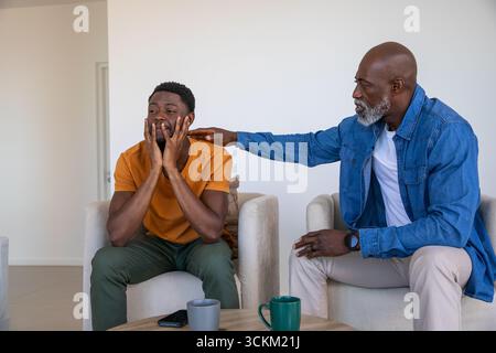 Père afro-américain et fils adultes se consolant dans le salon avec tasses, smartphone Banque D'Images