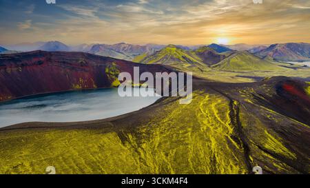 Un lac de cratère volcanique aux murs brun rougeâtre, entouré de mousse verte. Des montagnes volcaniques escarpées et un coucher de soleil doré sont visibles en arrière-plan Banque D'Images