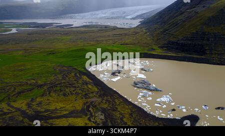 Un lagon glaciaire avec de l'eau brun laiteux et des icebergs flottants, entouré de terrain volcanique et de mousse verte, avec un glacier dans le fond brumeux. Banque D'Images