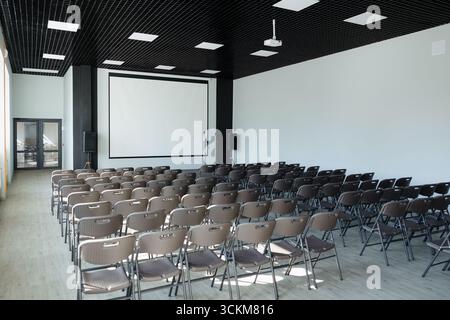 Des rangées de chaises brunes sont disposées dans une salle de conférence spacieuse et bien éclairée dotée d'un grand écran de projection. Banque D'Images