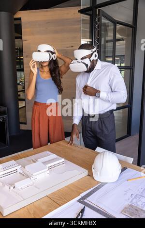 Divers collègues ajustant les casques vr, étudiant le modèle réduit et les plans sur la table dans la salle de réunion Banque D'Images