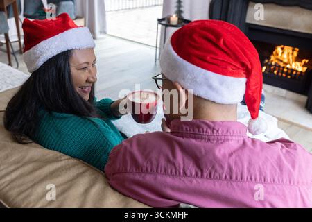 Couple diversifié dans les chapeaux de père noël sirotant des boissons de tasses rouges sur le canapé beige à côté de la cheminée Banque D'Images