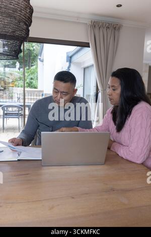 Couple senior diversifié examinant des documents imprimés et ordinateur portable argenté à la table à manger à la maison Banque D'Images