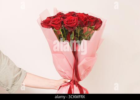 Bouquet de roses rouges enveloppé dans du papier rose. Fleurs de cadeau dans la main de la femme. Cadeau romantique pour la célébration et l'amour. Banque D'Images
