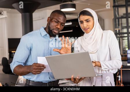 Divers collègues examinant l'ordinateur portable argenté, document papier dans la tenue d'affaires dans le bureau ouvert Banque D'Images
