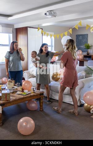 Diverses amies féminines se réunissant dans le salon autour de la table avec gâteau, cadeau enveloppé et ballons Banque D'Images