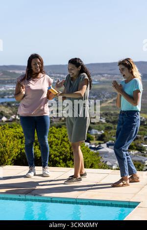 Diverses amies féminines pulvérisant avec un pistolet à eau sur la terrasse de la piscine carrelée près de la piscine surplombant la côte Banque D'Images