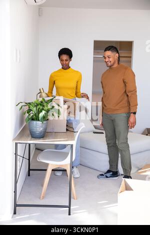 Couple diversifié déballer la boîte en carton à côté du bureau en bois dans le salon, enlever la lampe et les dossiers Banque D'Images