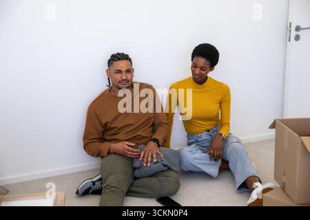 Couple diversifié assis sur le sol moquette clair dans la nouvelle pièce à la maison au milieu de boîtes déballées et smartphone Banque D'Images