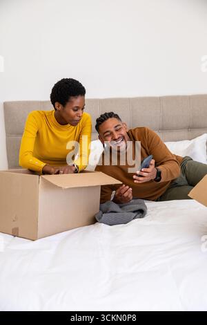 Couple diversifié déballer la boîte en carton sur le lit à la maison, homme riant au smartphone près du pull gris Banque D'Images