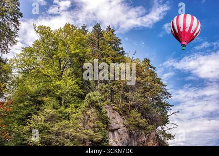 Une montgolfière rouge et blanche sur fond de hautes montagnes des Alpes avec forêt verte sous ciel nuageux bleu. Banque D'Images