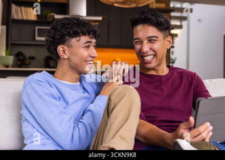 Deux amis masculins riant et partageant la tablette sur le canapé de couleur claire dans le salon Banque D'Images