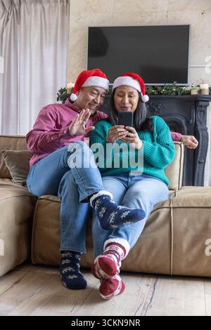 Couple senior assis sur le canapé dans les chapeaux de père noël dans le salon avec guirlande à l'aide d'un smartphone Banque D'Images
