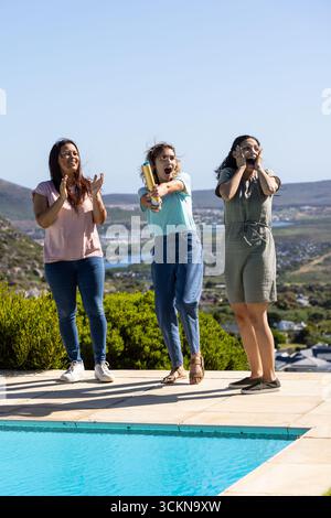 Diverses amies se tenant debout sur le patio près des arbustes de la piscine et éclatant des confettis dorés Banque D'Images