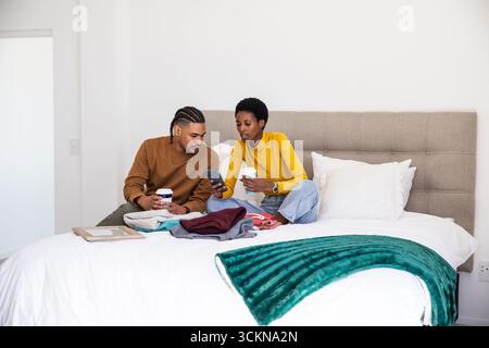 Couple diversifié assis sur le lit tenant des tasses à café et smartphone tout en triant les vêtements pliés Banque D'Images