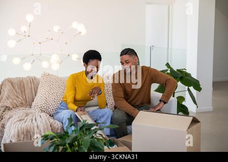 Couple diversifié déballer le cadre photo sur le canapé sous le lustre dans le salon au milieu des boîtes et de la plante Banque D'Images