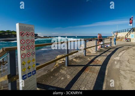 Panneau fournissant des consignes de sécurité près d'un sentier de bord de mer avec les gens appréciant le paysage côtier et la lumière du soleil, Clovelly Bay, Clovelly, Sydney Banque D'Images