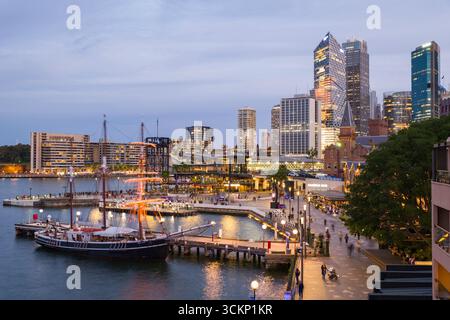 Vue sur une promenade animée du côté du port avec un voilier historique et un paysage urbain moderne, illuminé au crépuscule, The Rocks, Sydney Banque D'Images
