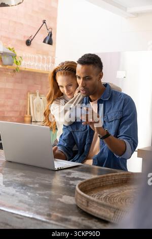 Couple diversifié embrassant tout en utilisant un ordinateur portable, smartphone sur la table de comptoir avec plateau en osier Banque D'Images