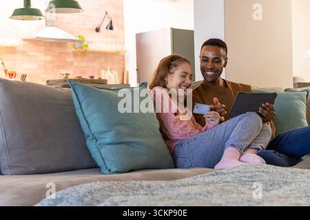 Couple diversifié s'appuyant sur un canapé dans un appartement moderne à l'aide d'une tablette et d'une carte de crédit, espace de copie Banque D'Images