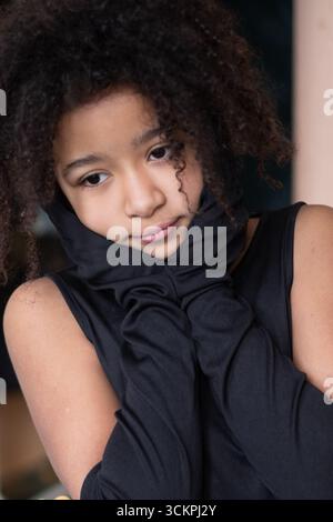 Fille afro-américaine aux cheveux bouclés pose de manière ludique avec ses mains sur ses joues, portant une tenue noire élégante, capturant un moment d'innocence A. Banque D'Images
