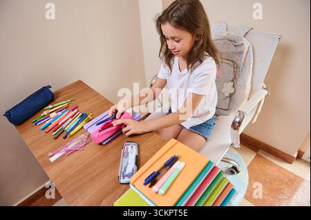 Une fille organise ses fournitures scolaires, y compris des stylos et des cahiers, sur un bureau à la maison. La configuration comprend divers outils colorés, favorisant l'apprentissage d'un Banque D'Images