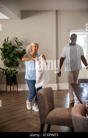 Couple diversifié debout dans le salon et la salle à manger tenant chemise blanche près de la plante en pot par porte Banque D'Images