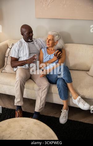 Couple senior diversifié assis sur le canapé velours côtelé à la maison discutant sur smartphone près de la table en marbre Banque D'Images