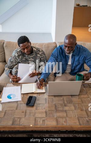 Père afro-américain et fils examinant les cartes avec lui en uniforme de camouflage sur le canapé Banque D'Images