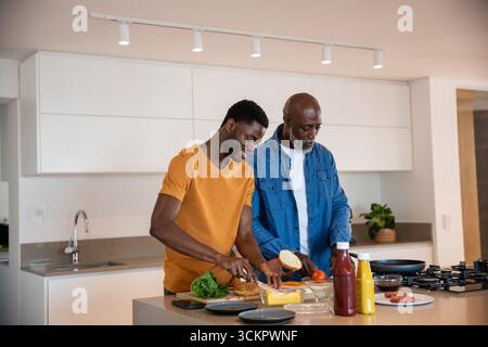 Père afro-américain et fils tranchant du pain et du fromage dans la cuisine sur une planche à découper Banque D'Images