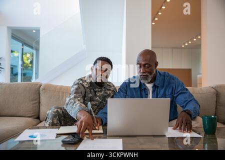 Père afro-américain et son fils en uniforme de camouflage à la maison examinant les cartes sur ordinateur portable Banque D'Images