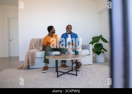 Père afro-américain et fils discutant sur tablette, tasses à café dans le salon Banque D'Images