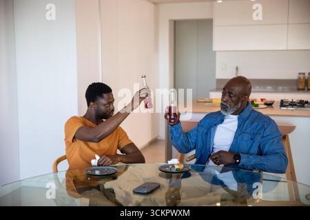 Père afro-américain et fils clinquant des bouteilles de boisson rouge à la table de la cuisine à la maison Banque D'Images
