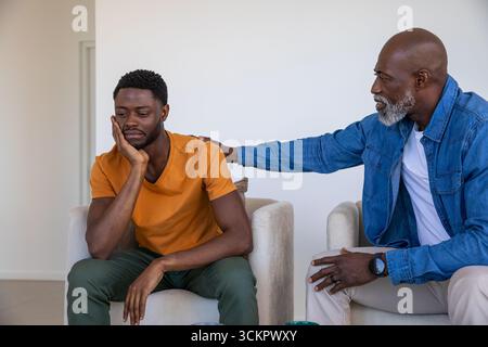 Père afro-américain réconfortant son fils inquiet assis dans le salon avec tasse à café Banque D'Images