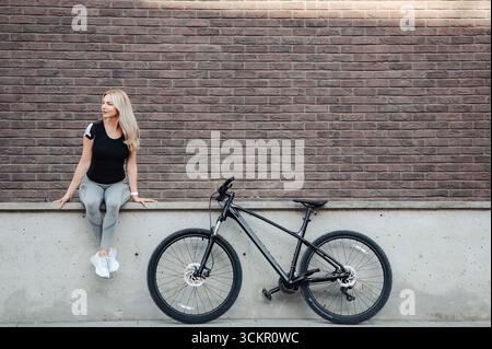Femme blonde mignonne se relaxant avec vélo par mur de brique après avoir roulé en plein air Banque D'Images