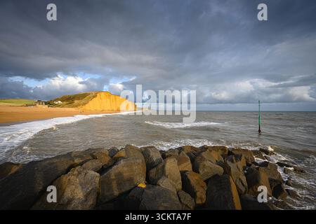 West Bay, Bridport, Dorset, Royaume-Uni. 12 septembre 2025. Météo britannique : les célèbres falaises de la station balnéaire de West Bay brillent orange alors qu'elles attrapent le soleil sur fond de nuages épais lors d'un après-midi de fortes averses et de périodes ensoleillées. Crédit : Celia McMahon/Alamy Live News Banque D'Images