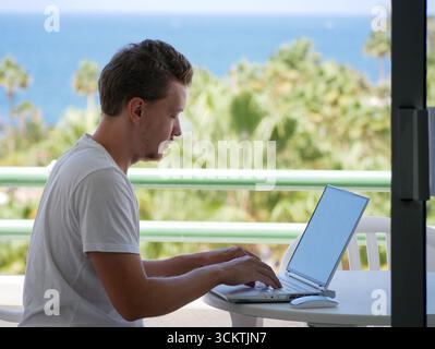 Un jeune homme travaille à distance sur ordinateur portable tout en étant assis à l'extérieur dans un endroit tropical. Thèmes du travail freelance, mode de vie nomade numérique, balan travail-vie Banque D'Images