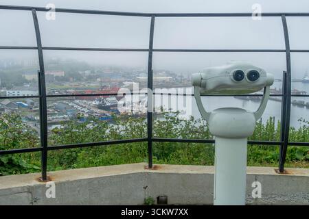 Visionneuse jumelle à pièces sur une colline surplombant le port brumeux et la ville de Korsakov, île de Sakhaline, Russie, avec des navires et des bâtiments industriels Banque D'Images