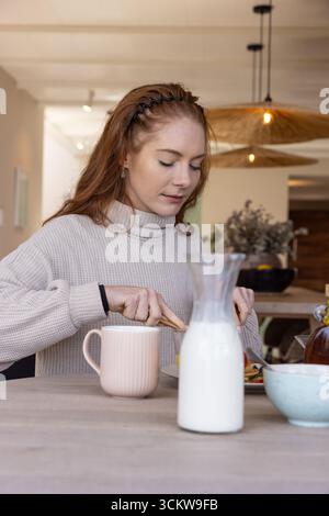 Femme coupant la nourriture sur l'assiette à la table à manger à la maison avec carafe à lait et tasse Banque D'Images