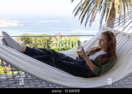 Femme allongée sur un hamac blanc à la terrasse du bord de mer dans une surchemise verte et tenant un smartphone Banque D'Images