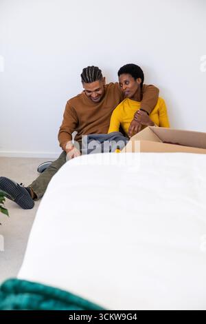 Couple diversifié assis sur la moquette dans la chambre déballant la boîte en carton avec couverture grise Banque D'Images