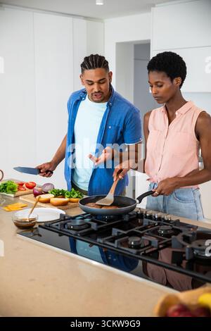 Couple diversifié préparant des hamburgers sur l'îlot de cuisine à l'aide d'une poêle, d'une spatule et d'un couteau de chef Banque D'Images