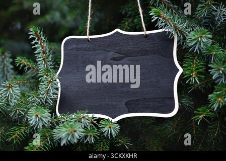 Tableau noir décoratif avec un cadre en bois, accroché à la ficelle parmi les branches de conifères verts. Banque D'Images